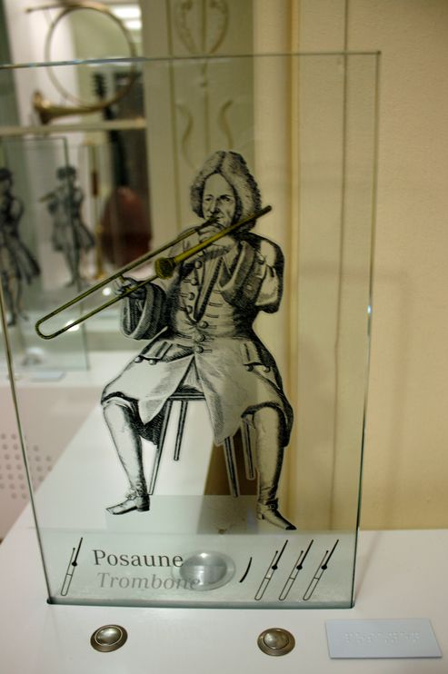 Glasscheibe mit Illustration (Musiker mit historischer Violine) und zwei Knöpfe, Braillebeschriftung