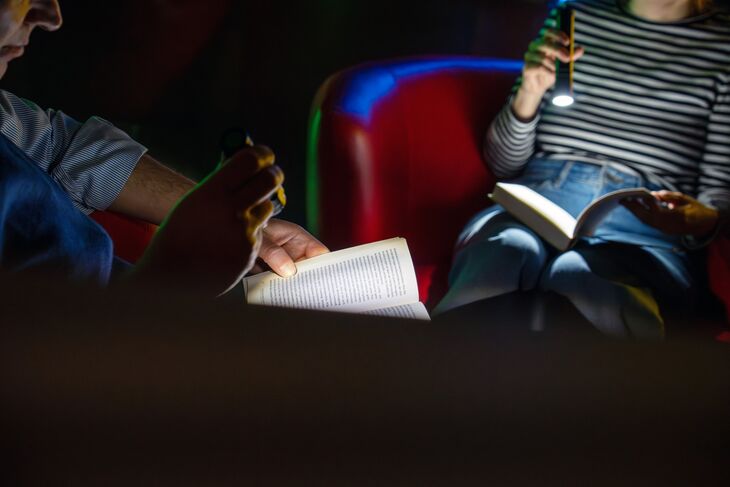 Zwei Menschen, die sich im Dunkeln gegenübersitzen, halten ein Buch auf ihrem Schoß und einen Taschenlampe in ihrer Hand