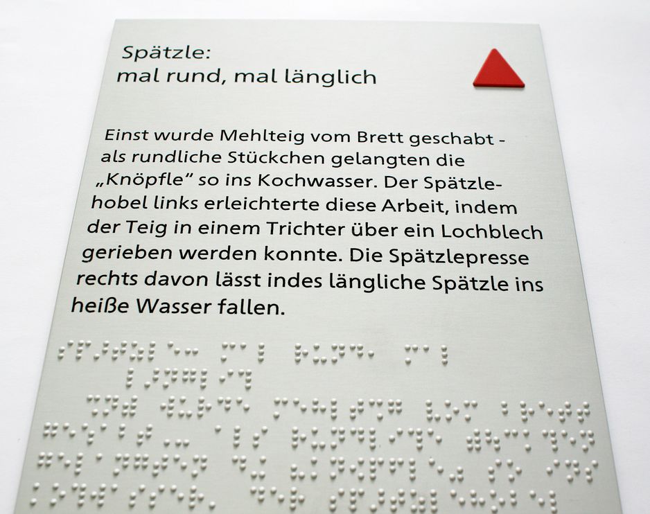 Aluminiumschild Hochformat mit Text in Schwarzdruck und Brailleschrift, rechts oben ein rotes taktiles Dreieck