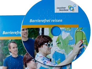 CD-Hülle auf der eine CD liegt: Label "Barrierefrei reisen" zeigt Frau mit Brille, die Landkarte ertastet